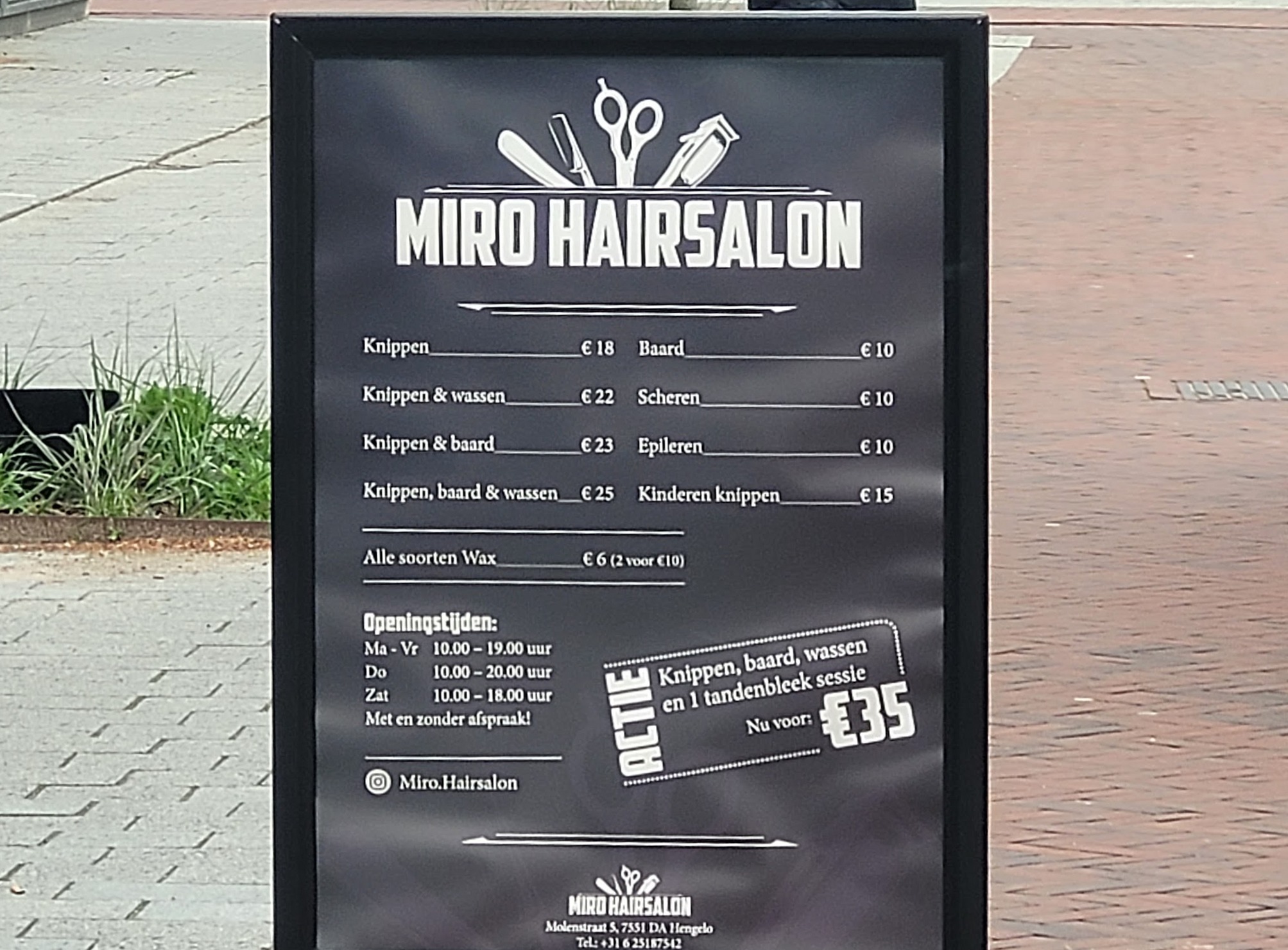 Het zwarte prijsbord van Miro Hairsalon op de stoep, met logo, prijzen, openingstijden en de actie